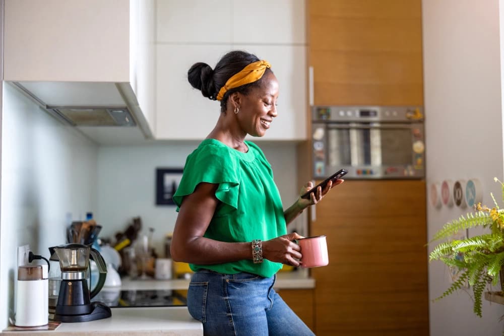 Woman smiling at her phone in a bright kitchen, coffee in hand